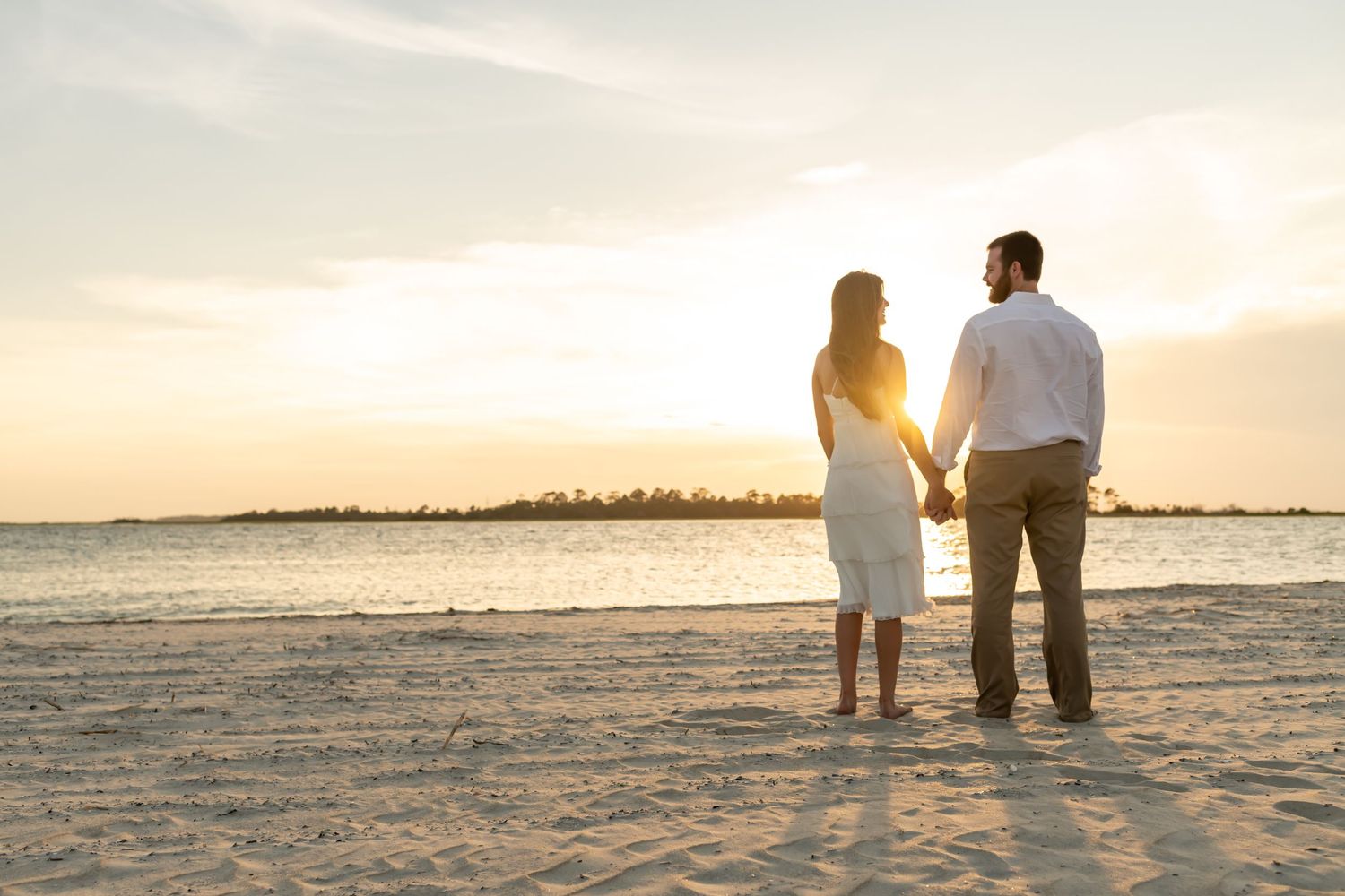 A couple stands together on a sandy beach at sunset, silhouetted against the golden sky and ocean horizon.