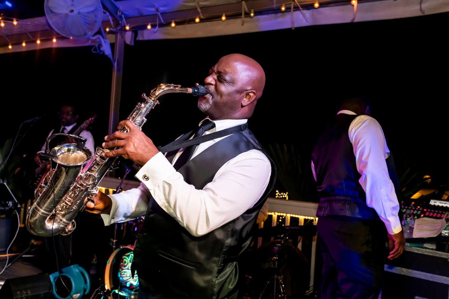 A saxophone player in a dark vest performs on stage with colorful lighting and decorations in the background.
