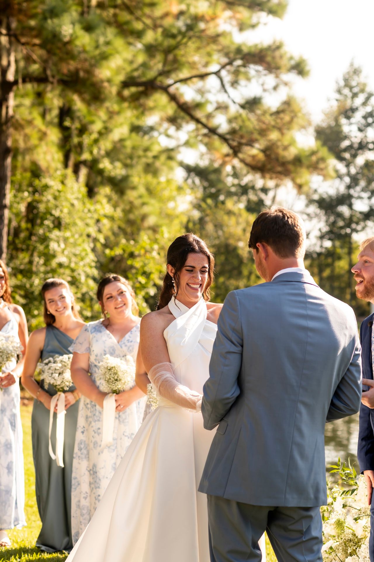 A wedding ceremony takes place outdoors in a sunlit forest setting with bridesmaids in elegant dresses watching the exchange.