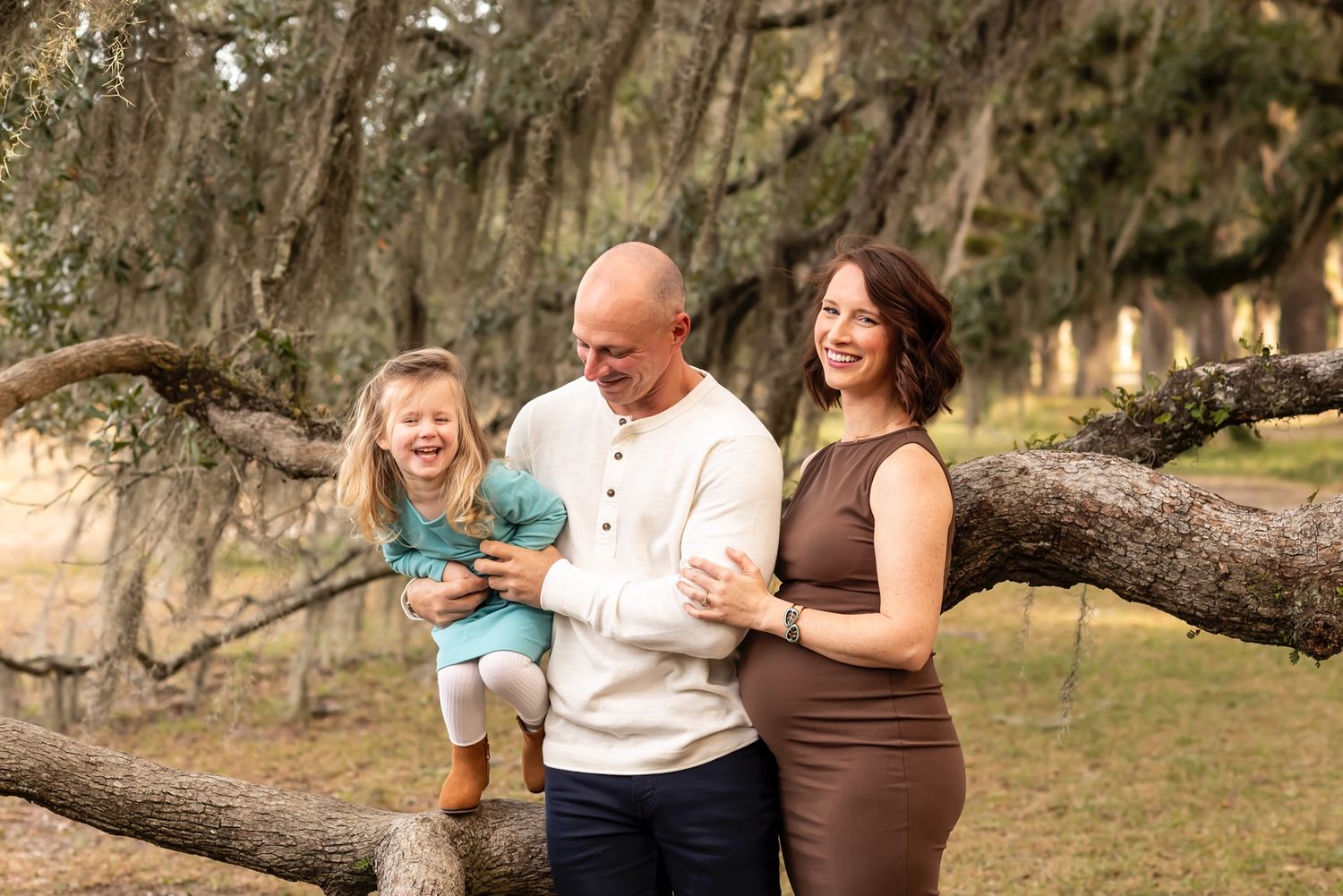 Three people stand together near a sweeping oak tree branch draped with Spanish moss on a sunny afternoon outdoors.
