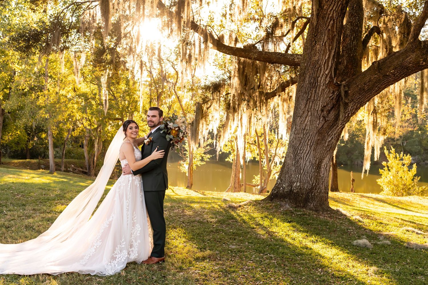 Two people embrace on their wedding day in a dreamy southern garden setting with Spanish moss and golden sunlight.
