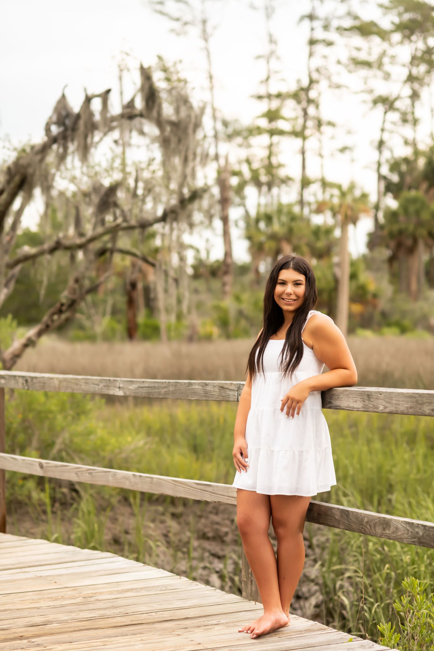 Barefoot woman in white sundress standing on wooden boardwalk near marsh and Spanish moss on summer day.