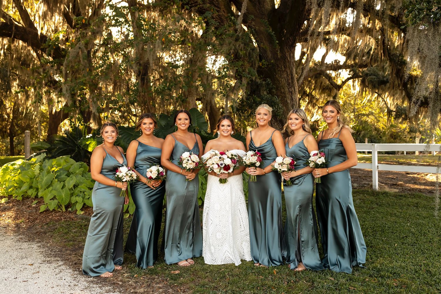 Wedding party in slate blue dresses poses together on a sunny day under oak trees at an outdoor venue.