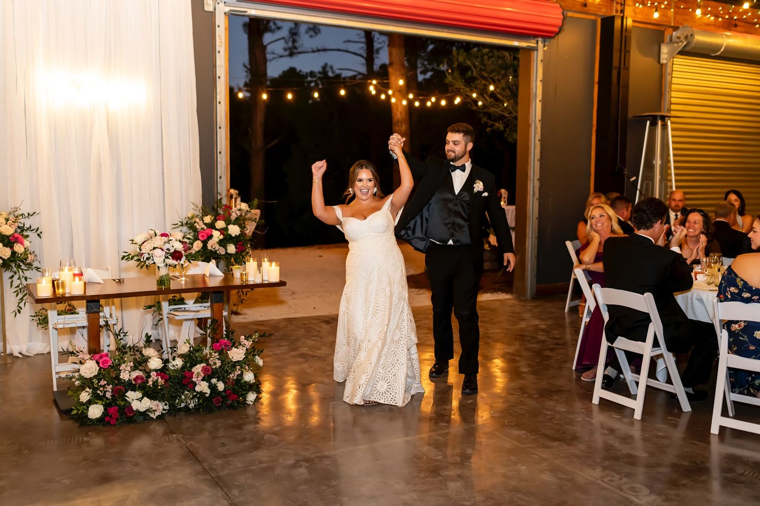 A couple makes their grand entrance at a wedding reception in an industrial venue decorated with string lights and flowers.