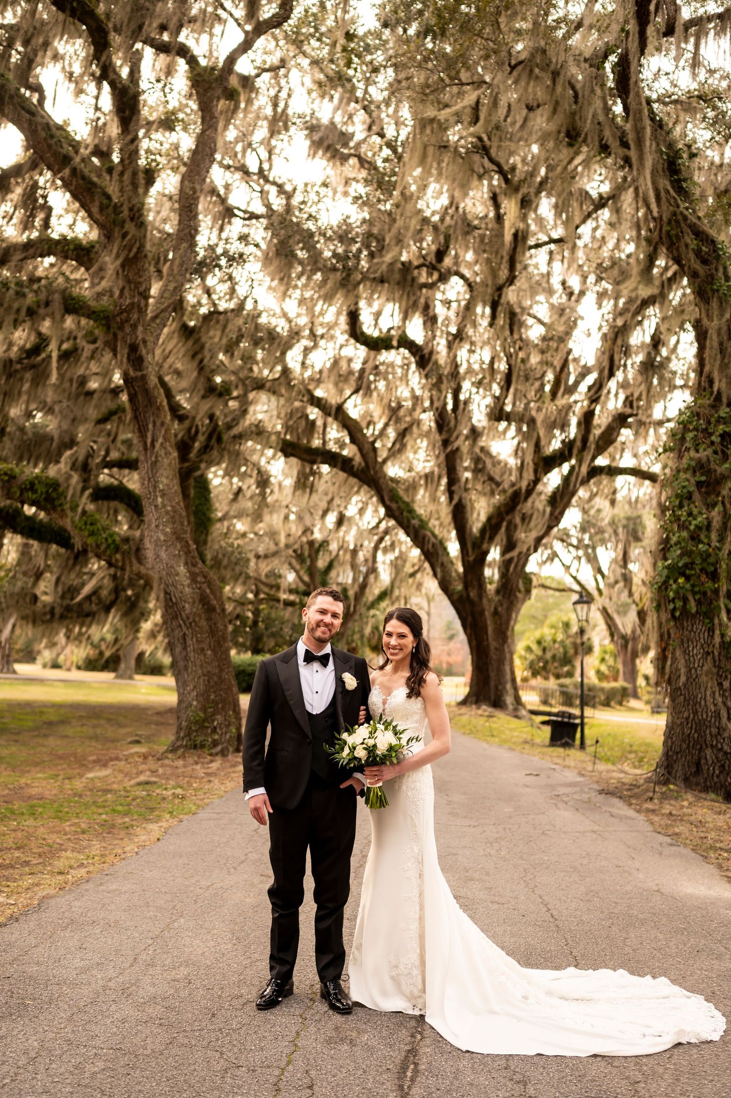 A couple stands on a pathway beneath Spanish moss draped oak trees at their wedding in savannah georgia.