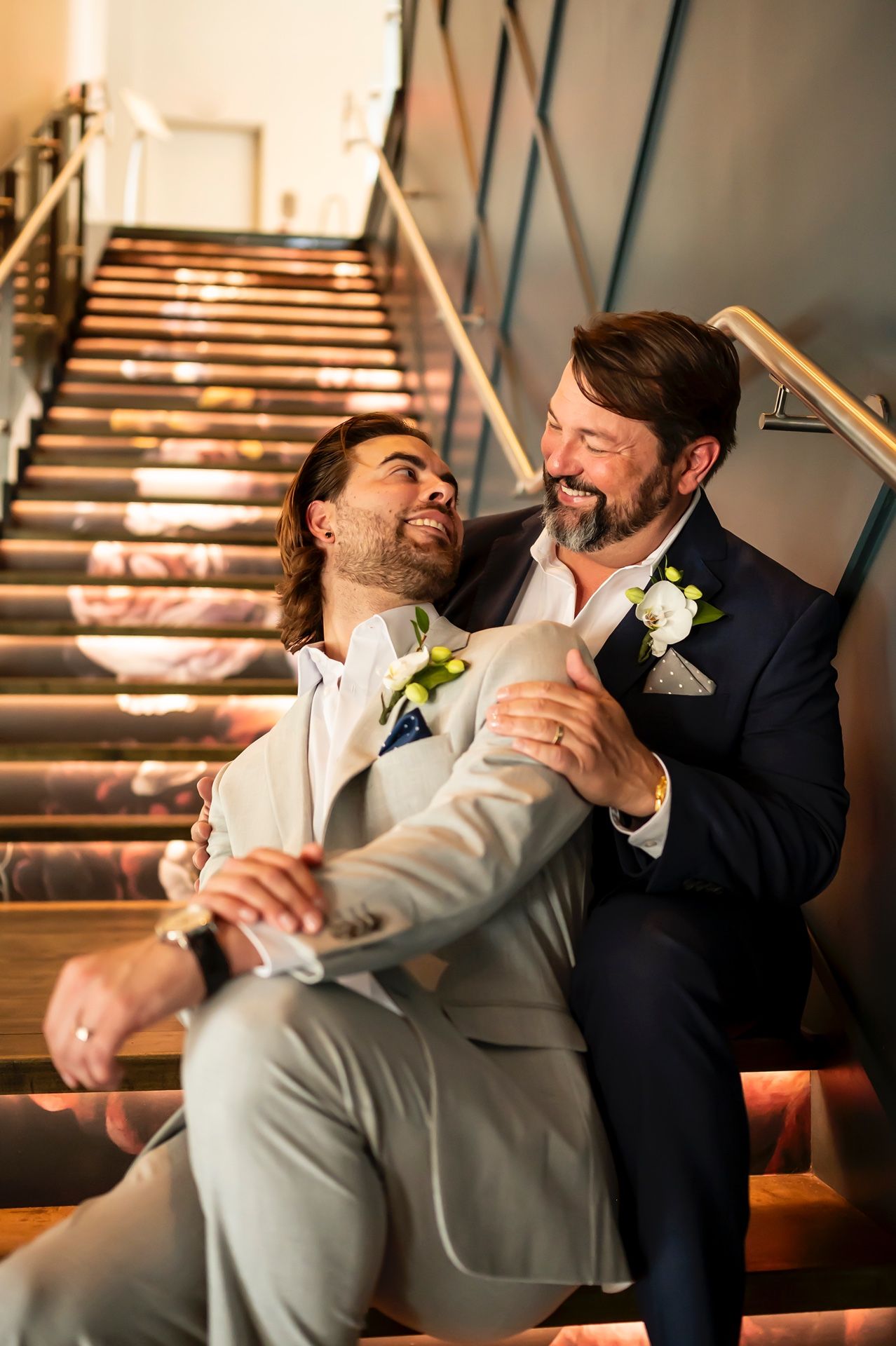 Two grooms share joyful laughter while sitting together on elegant wooden stairs at their wedding celebration.