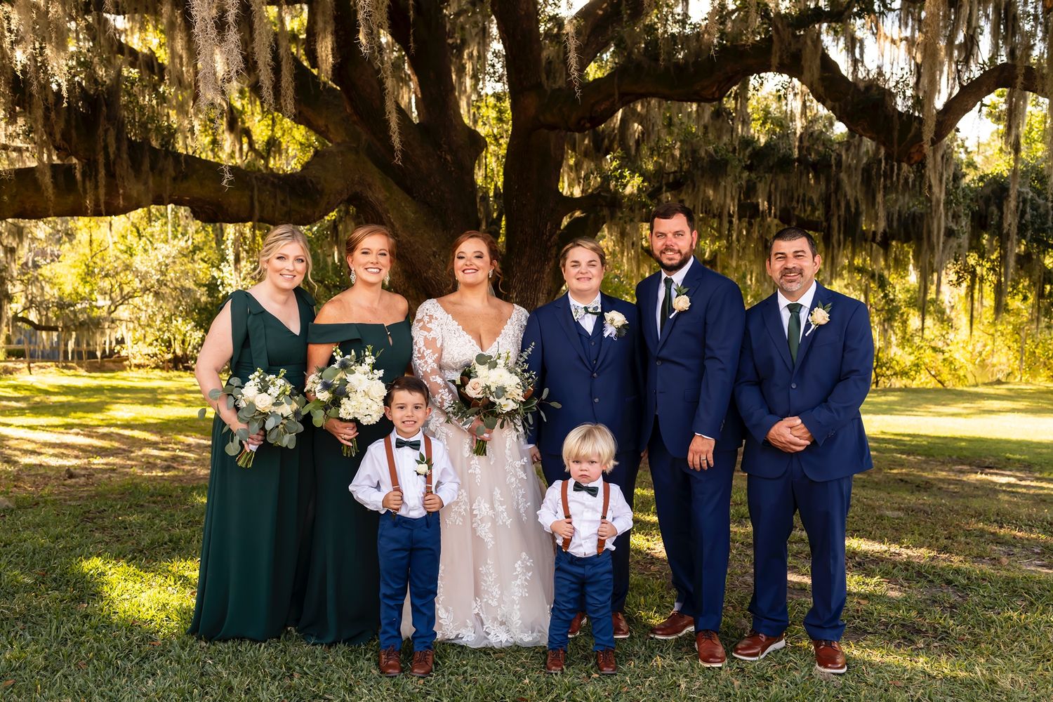 A wedding party poses outdoors under a large oak tree draped with Spanish moss in a sunlit garden setting.