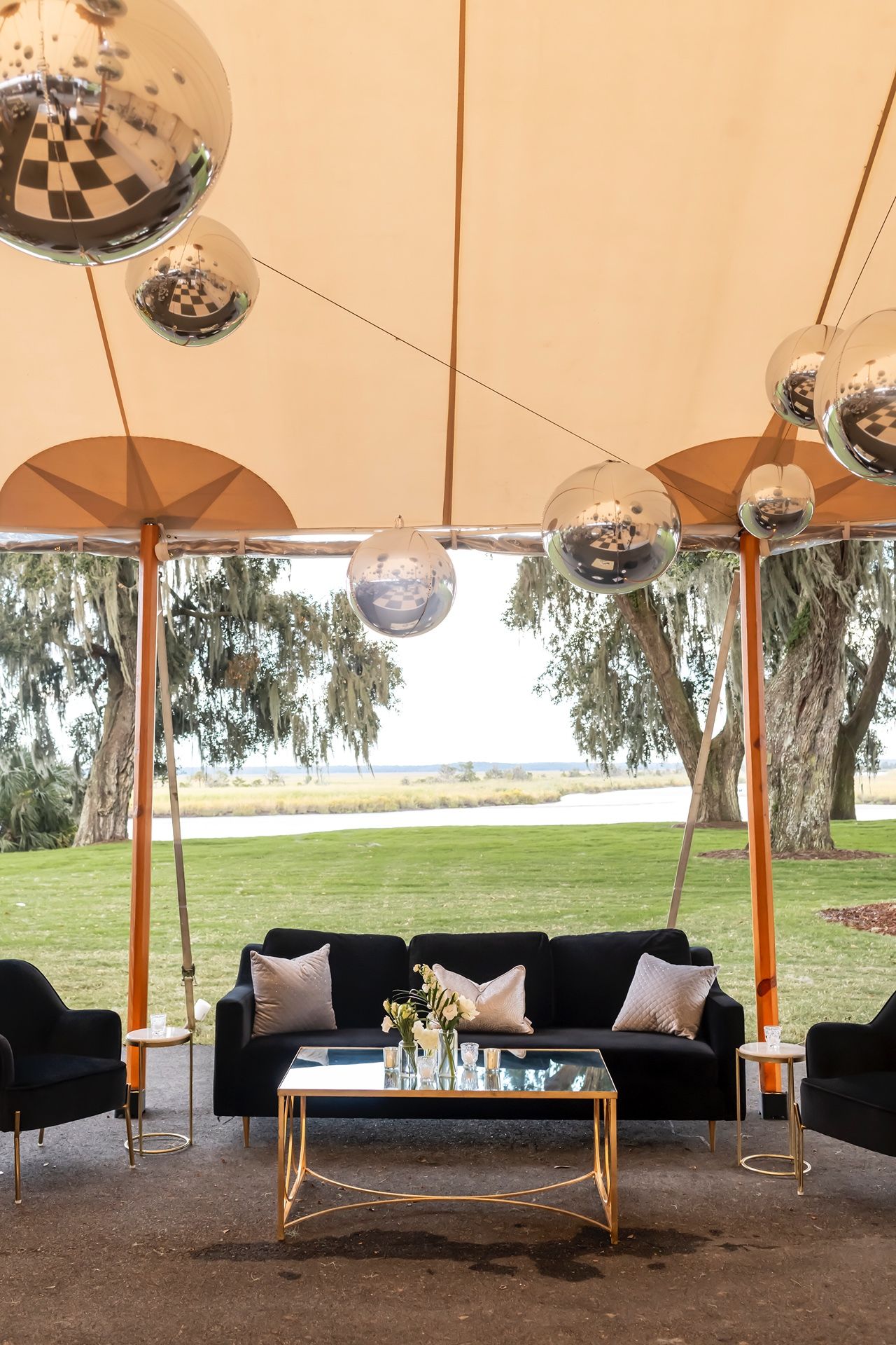 Luxurious outdoor lounge setup with disco balls hanging from tent canopy over black sofas and gold coffee table by waterfront.