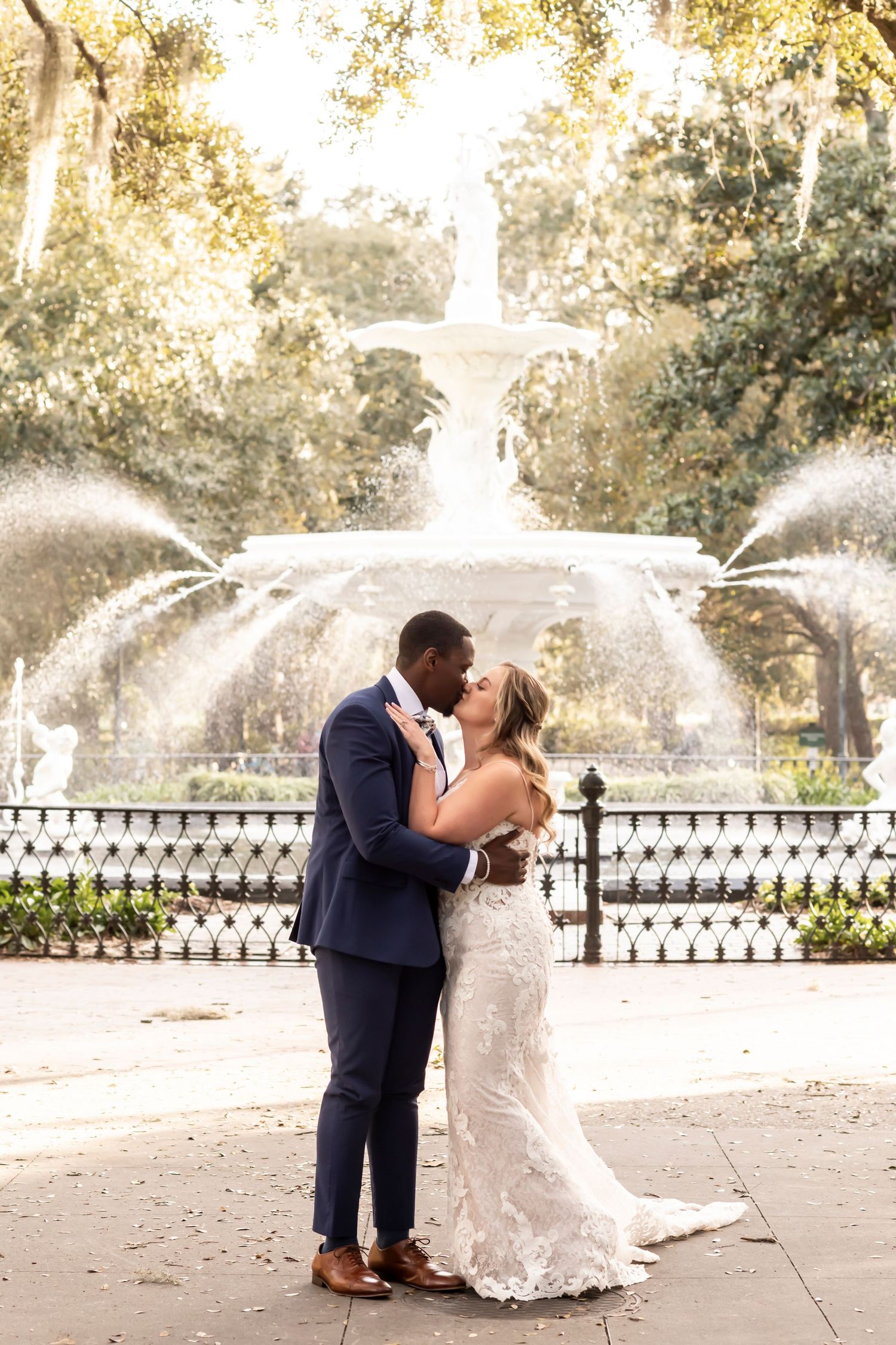 A romantic wedding couple embraces in front of a majestic tiered fountain in a scenic garden setting.