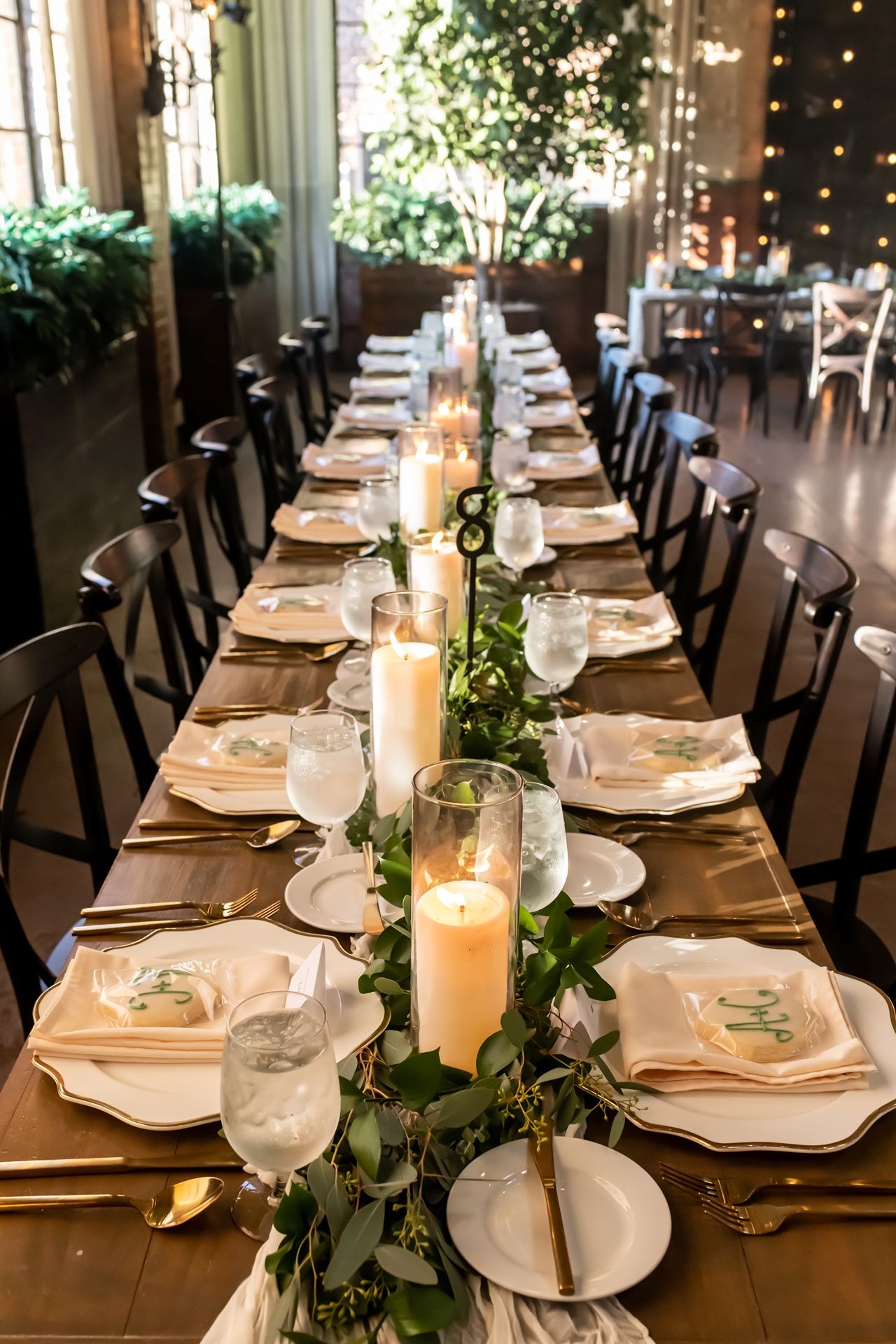 Elegant wedding reception table settings with white plates, black chairs, and greenery garlands illuminated by candlelight.