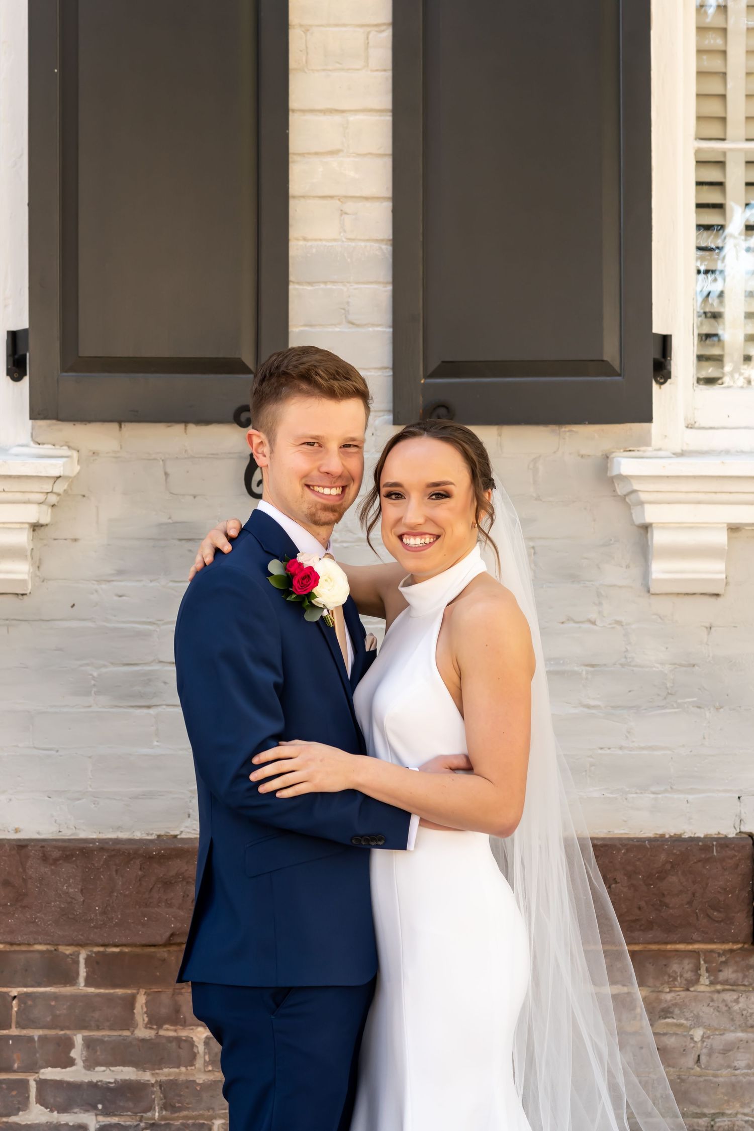 Wedding couple embraces against white brick wall with black shutters, featuring navy suit and sleek white gown with veil.