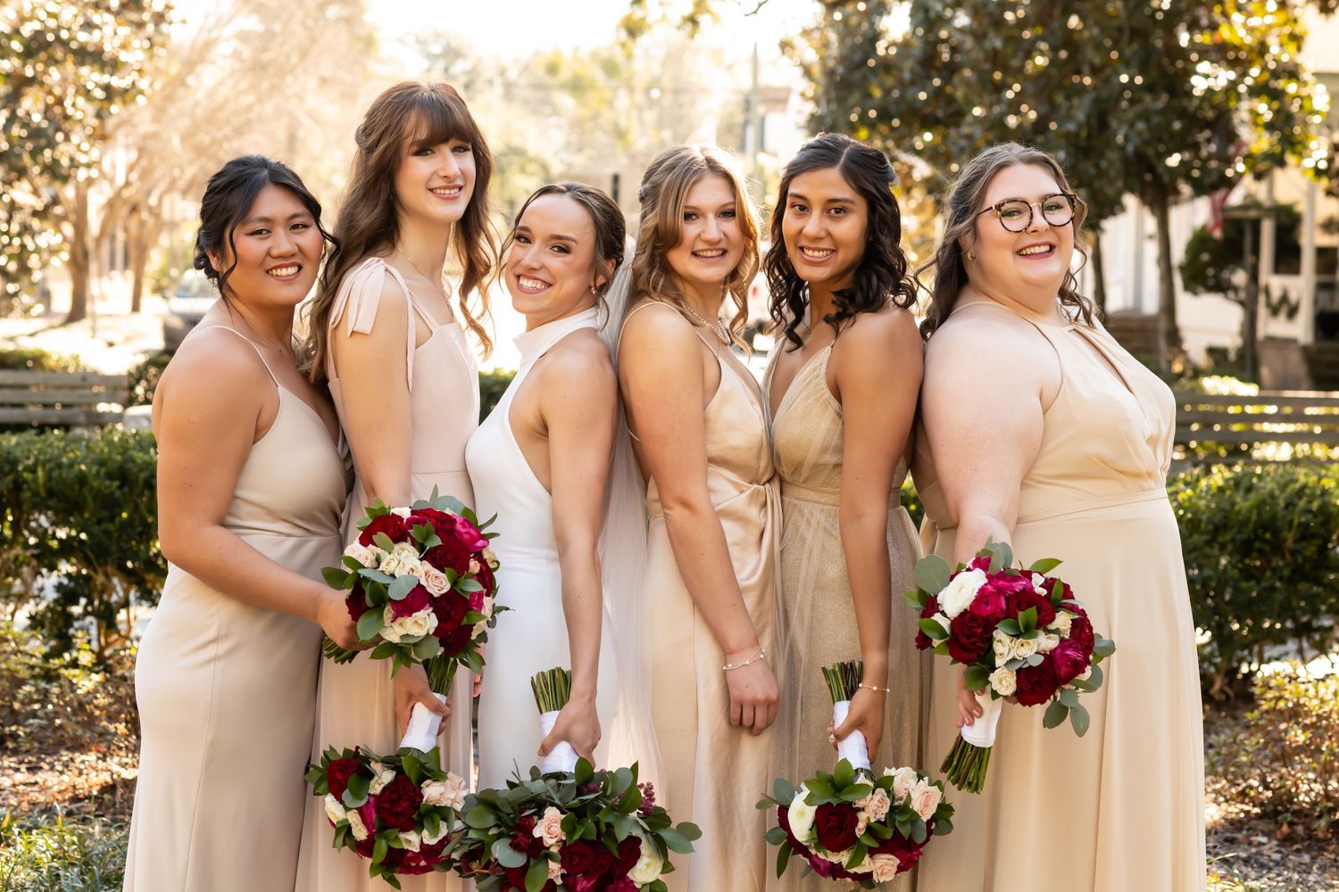 Bridesmaids wearing blush-colored dresses stand together smiling and laughing in a beautiful outdoor setting with fall foliage.