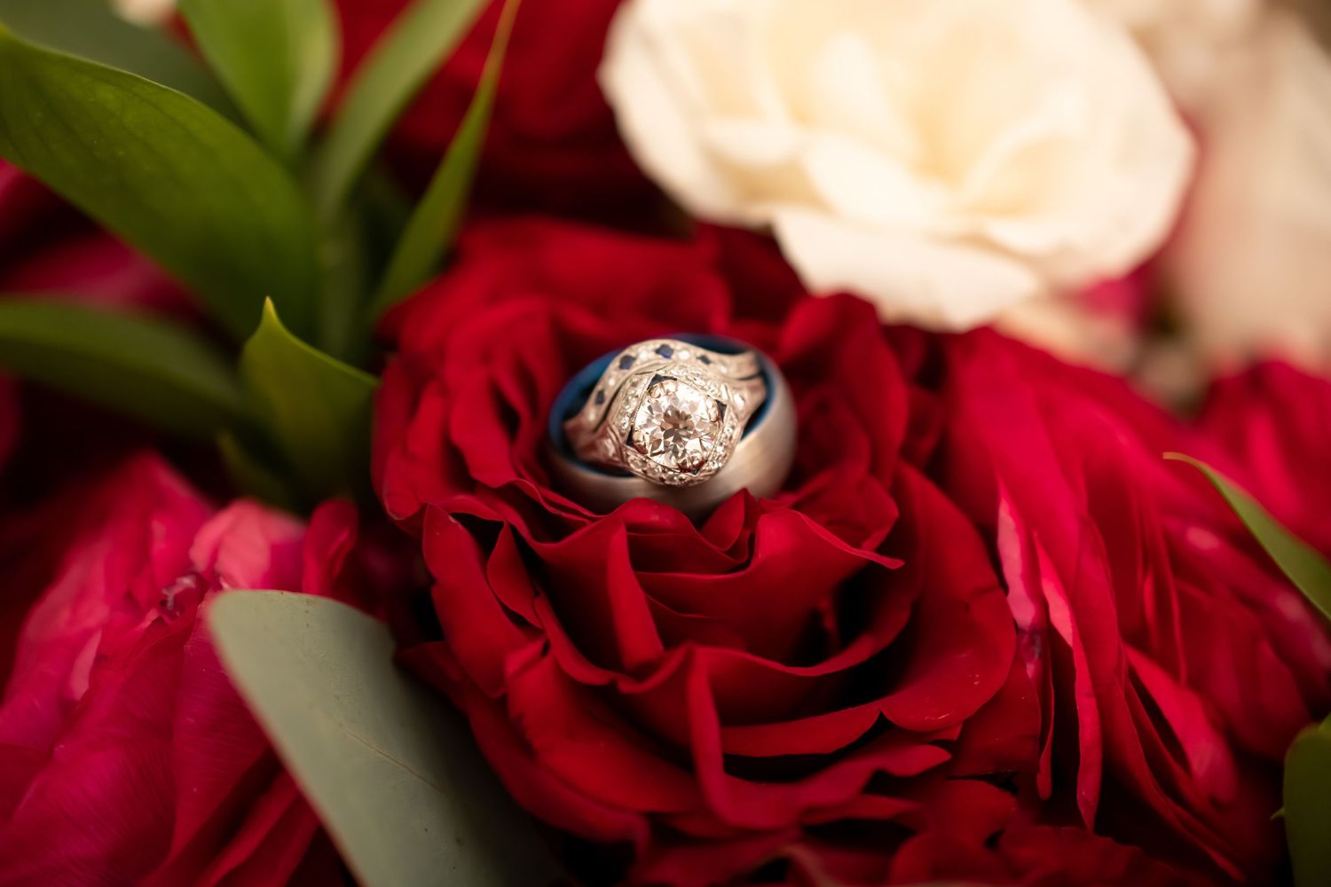 A diamond engagement ring nestles in the center of a vibrant red rose bloom among mixed floral arrangements.