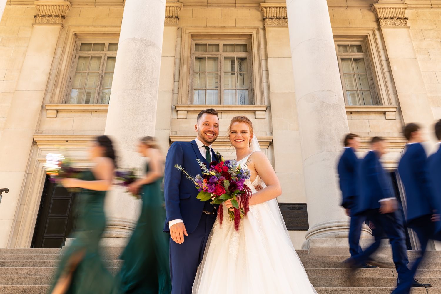 Motion-blurred guests walk past while newlyweds pose between white columns at a grand historic building entrance.