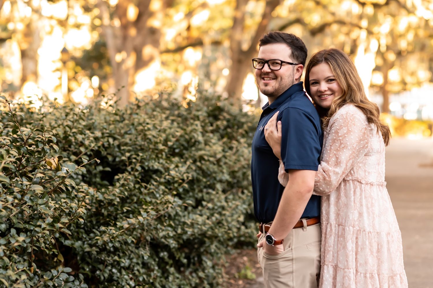 A couple shares an intimate moment while standing outdoors during golden hour among trees and greenery.