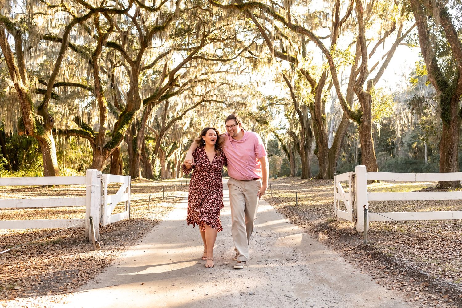 Two individuals walk together down a sunlit oak-lined path with Spanish moss and a white fence at sunset.