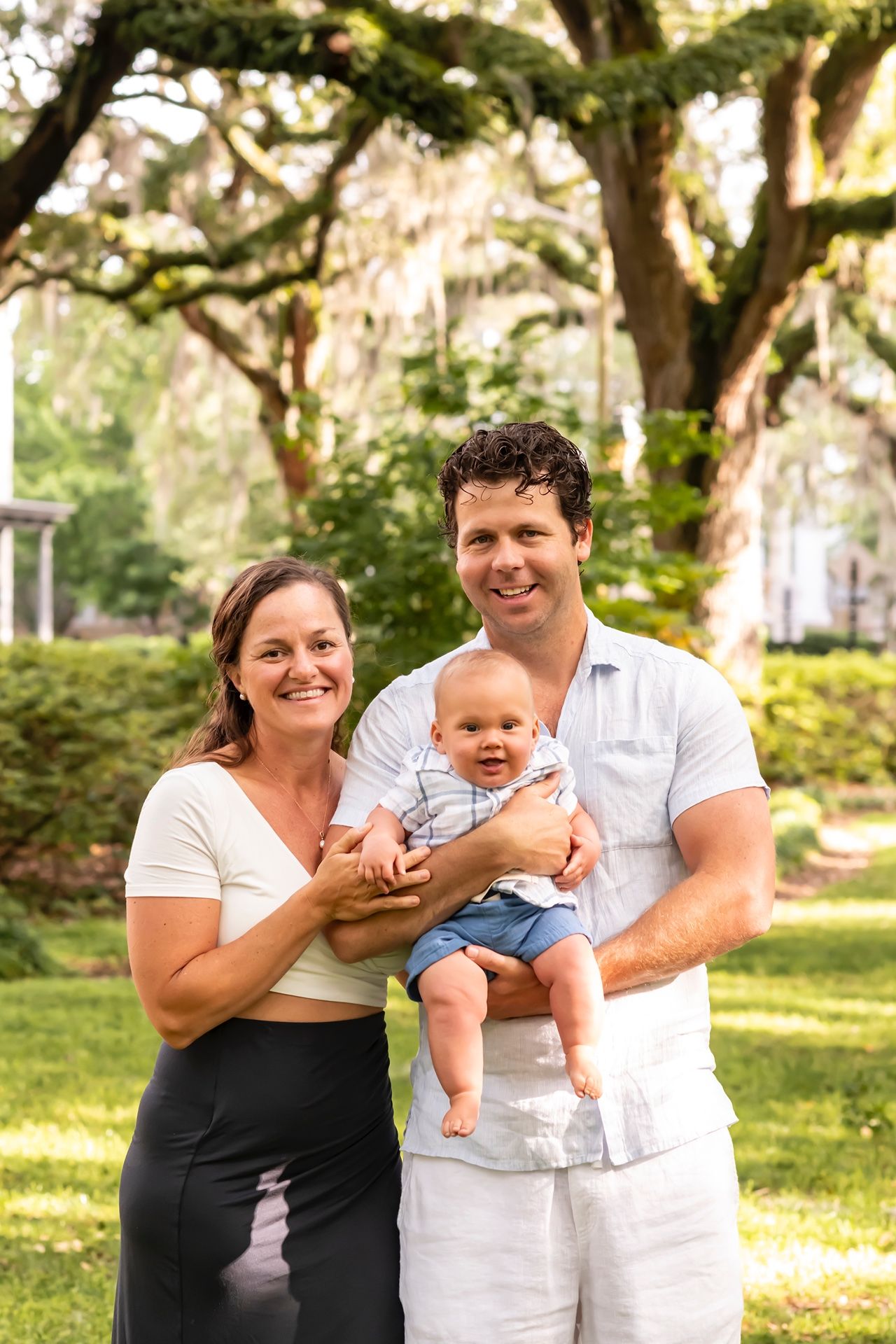 Family stands together outdoors under mossy oak trees in a sunny garden setting with infant.