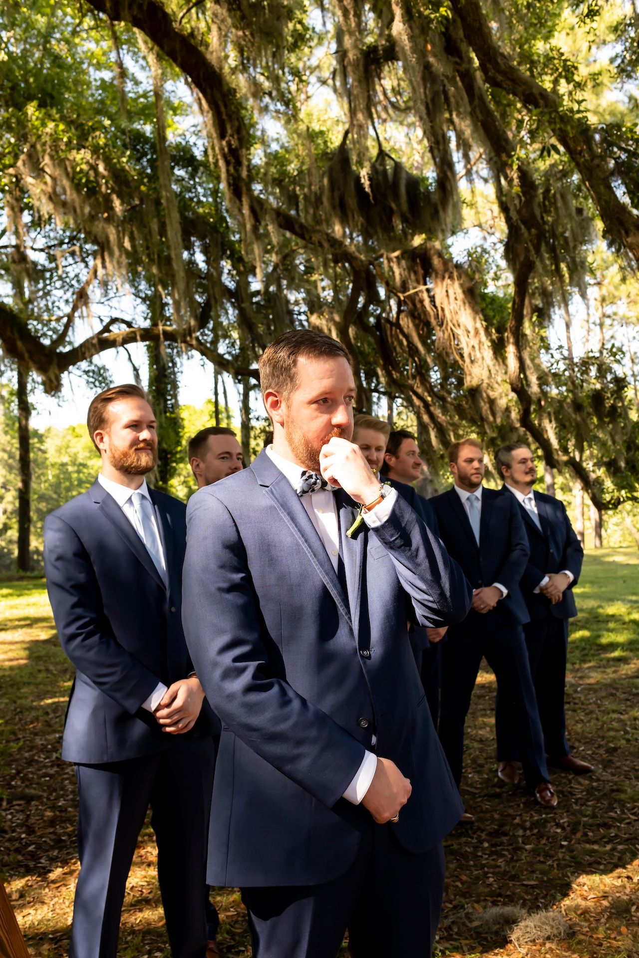 Groomsmen in navy suits stand beneath Spanish moss-draped oak trees at a Southern wedding ceremony.