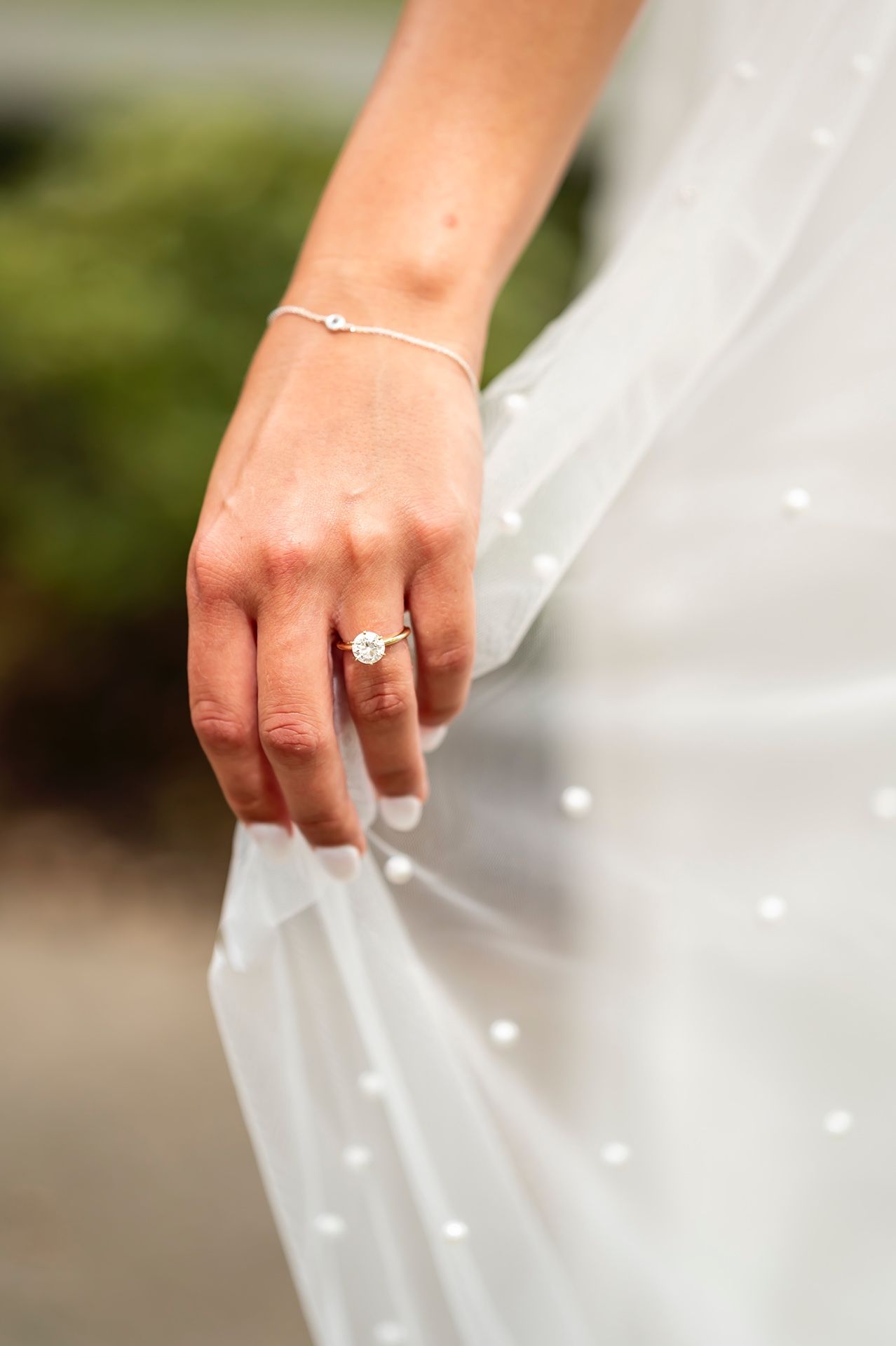 A delicate hand wearing an engagement ring and bracelet gently holds white tulle veil fabric with scattered pearl details.