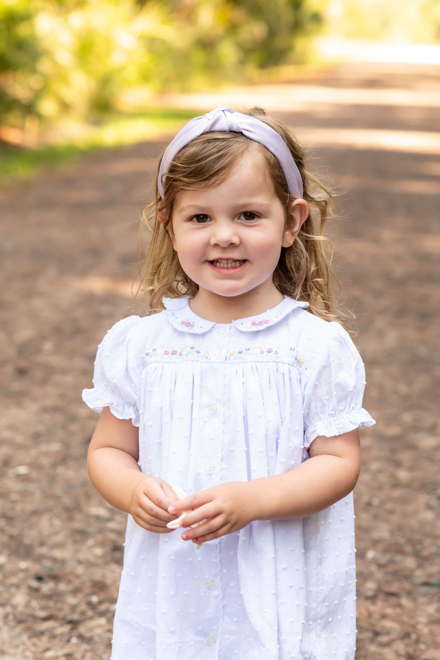 A young child in a white eyelet lace dress poses on a dirt path surrounded by sunlit greenery.