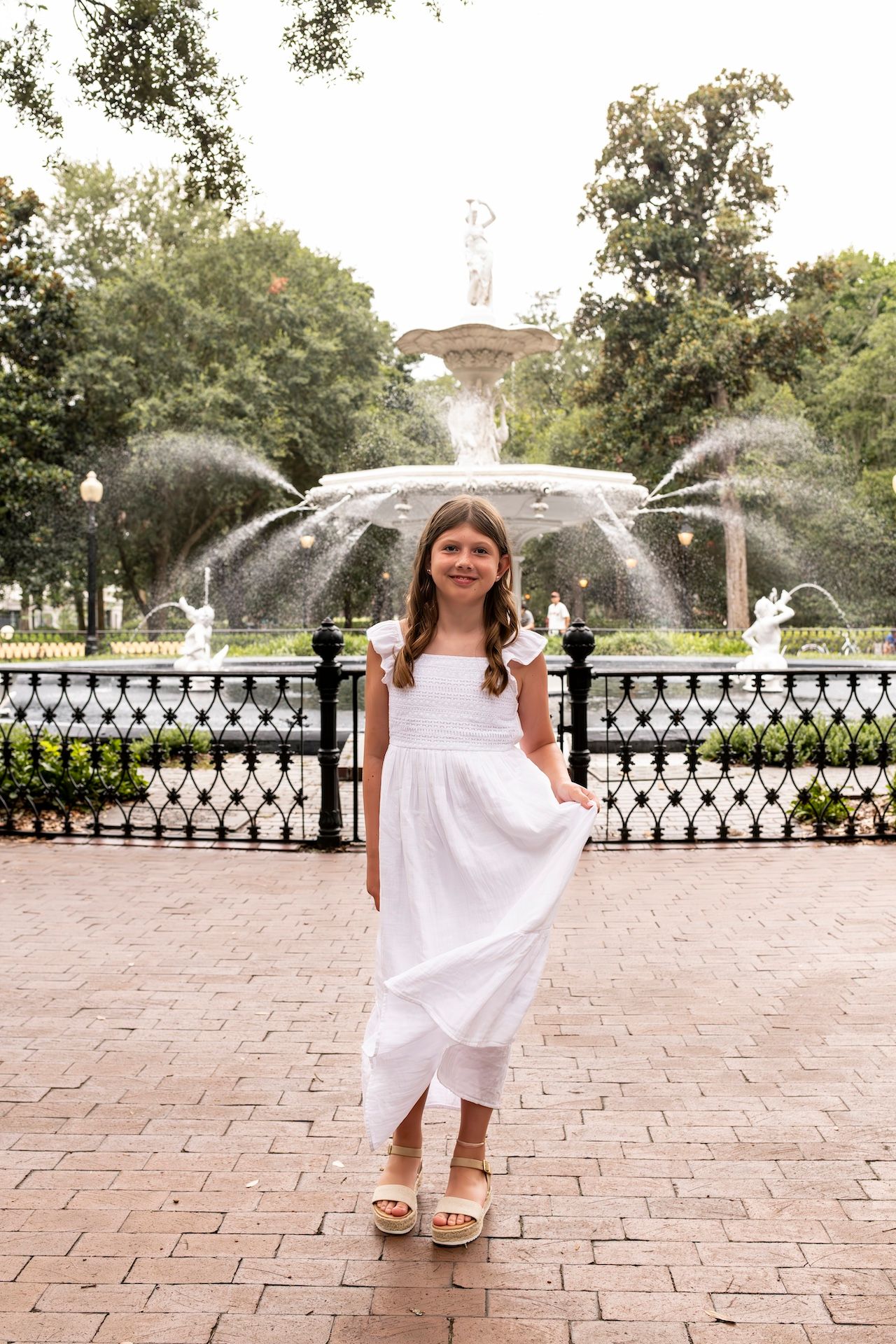 A young person in a white dress poses in front of an ornate fountain with water sprays.