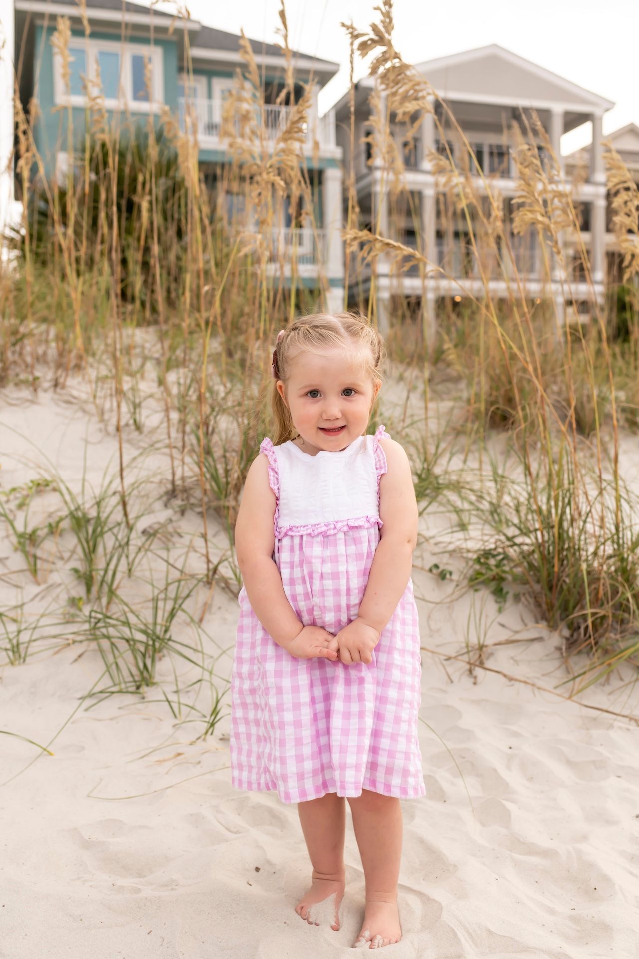 Pink gingham dress on sandy beach dunes with coastal homes and sea oats in background.