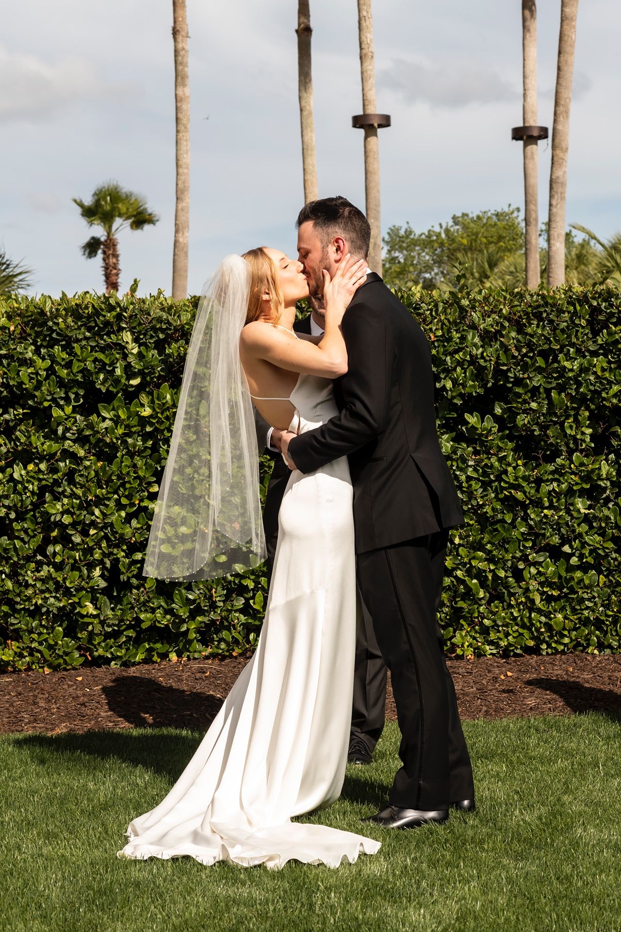 Newlyweds share an intimate moment in their wedding attire on a manicured lawn with palm trees and hedges in the background.