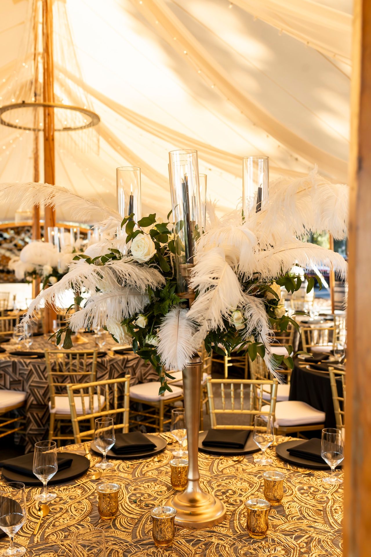 Elegant wedding reception table with gold accents, white feather centerpieces and geometric patterned tablecloth under tent lighting.