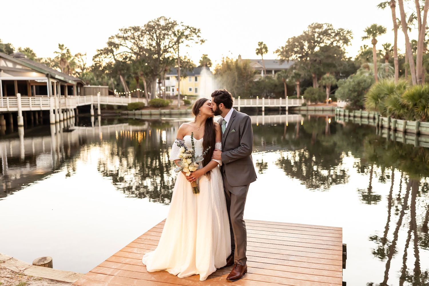 A couple shares a romantic moment on a waterfront dock at sunset with palm trees and buildings in the background.