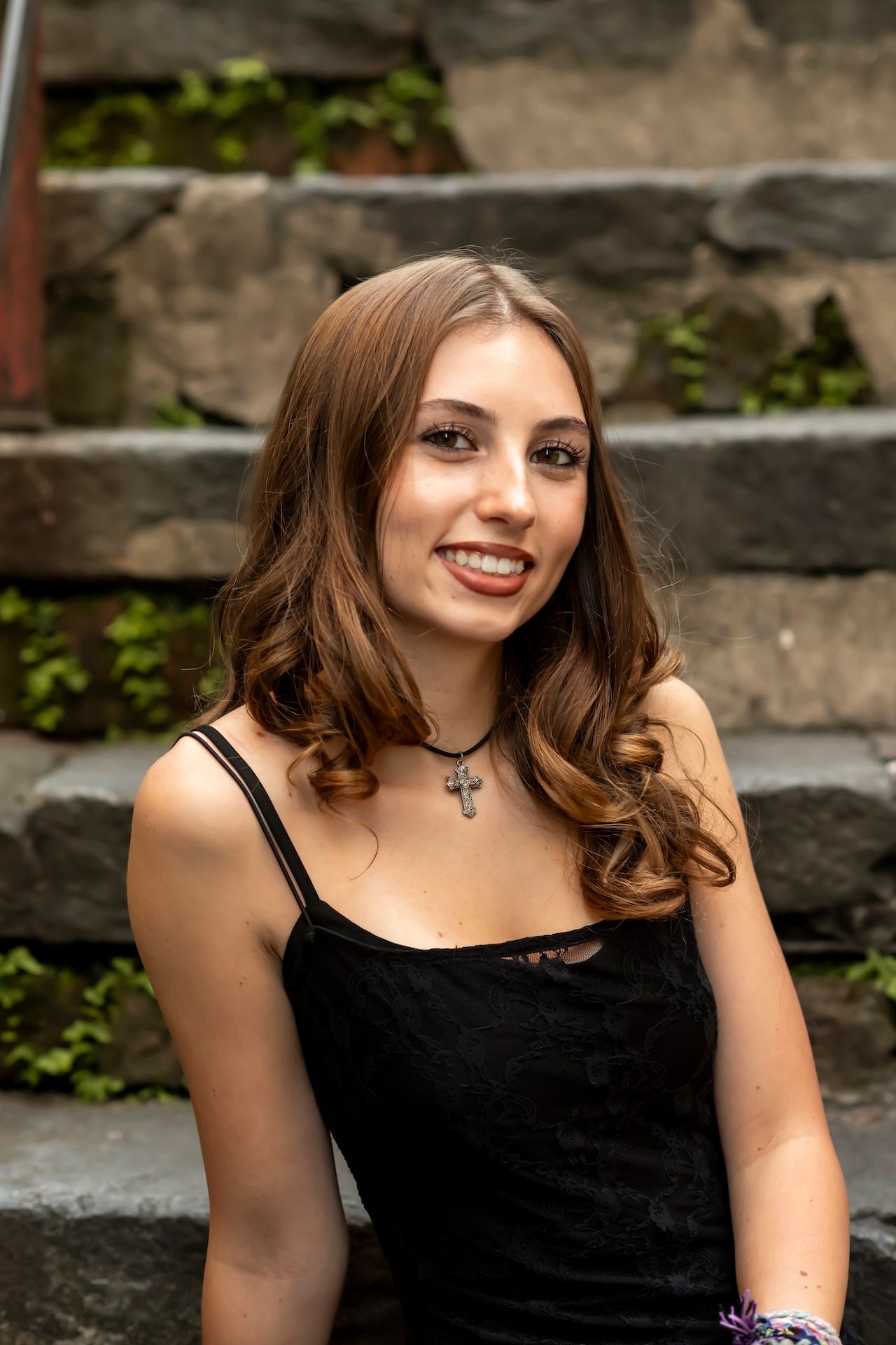 Portrait of someone in black top with long brown hair sitting on outdoor stone steps with greenery.