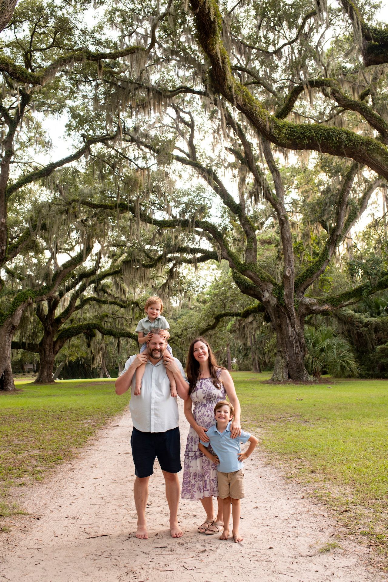 Family of four posing on a sandy path beneath sprawling moss-draped oak trees in a lush green park.