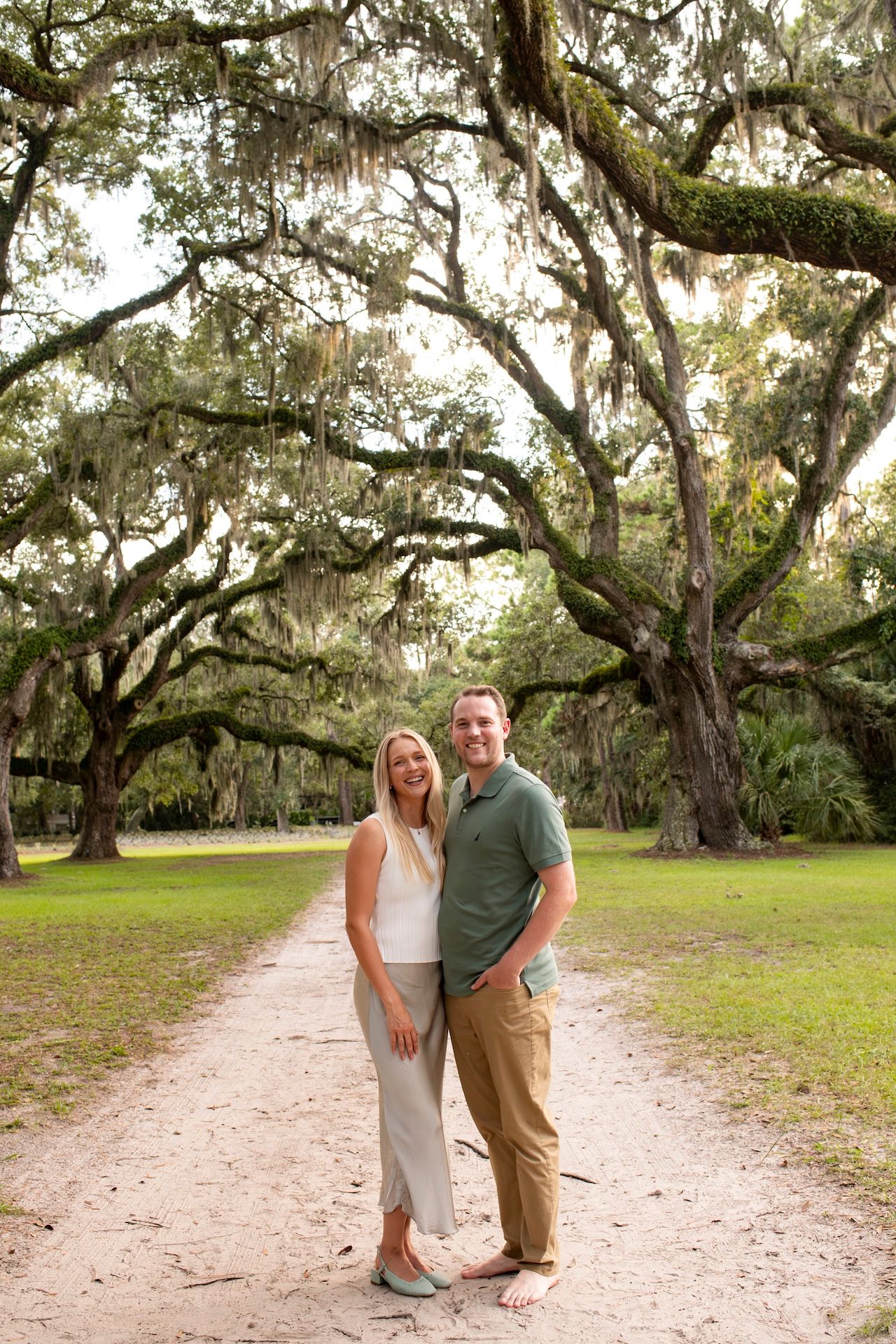 Smiling couple posing on a sandy path under large moss-draped oak trees in a scenic outdoor setting.