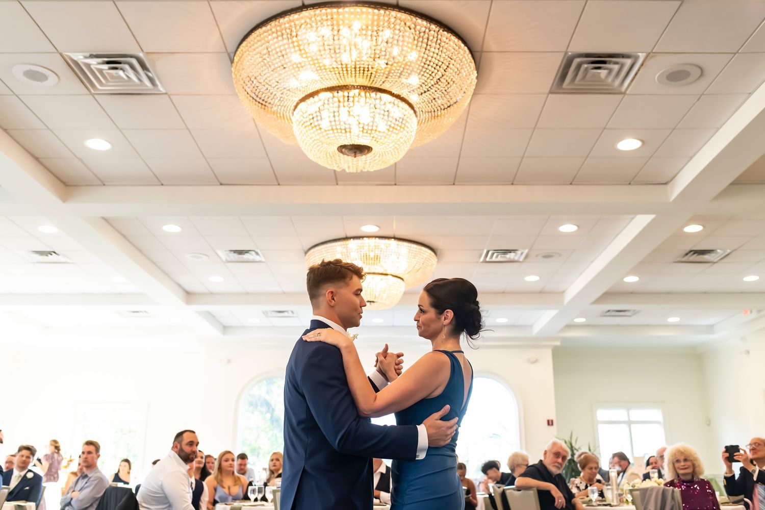 Two people share their first dance as newlyweds at an elegant indoor wedding reception with crystal chandeliers.