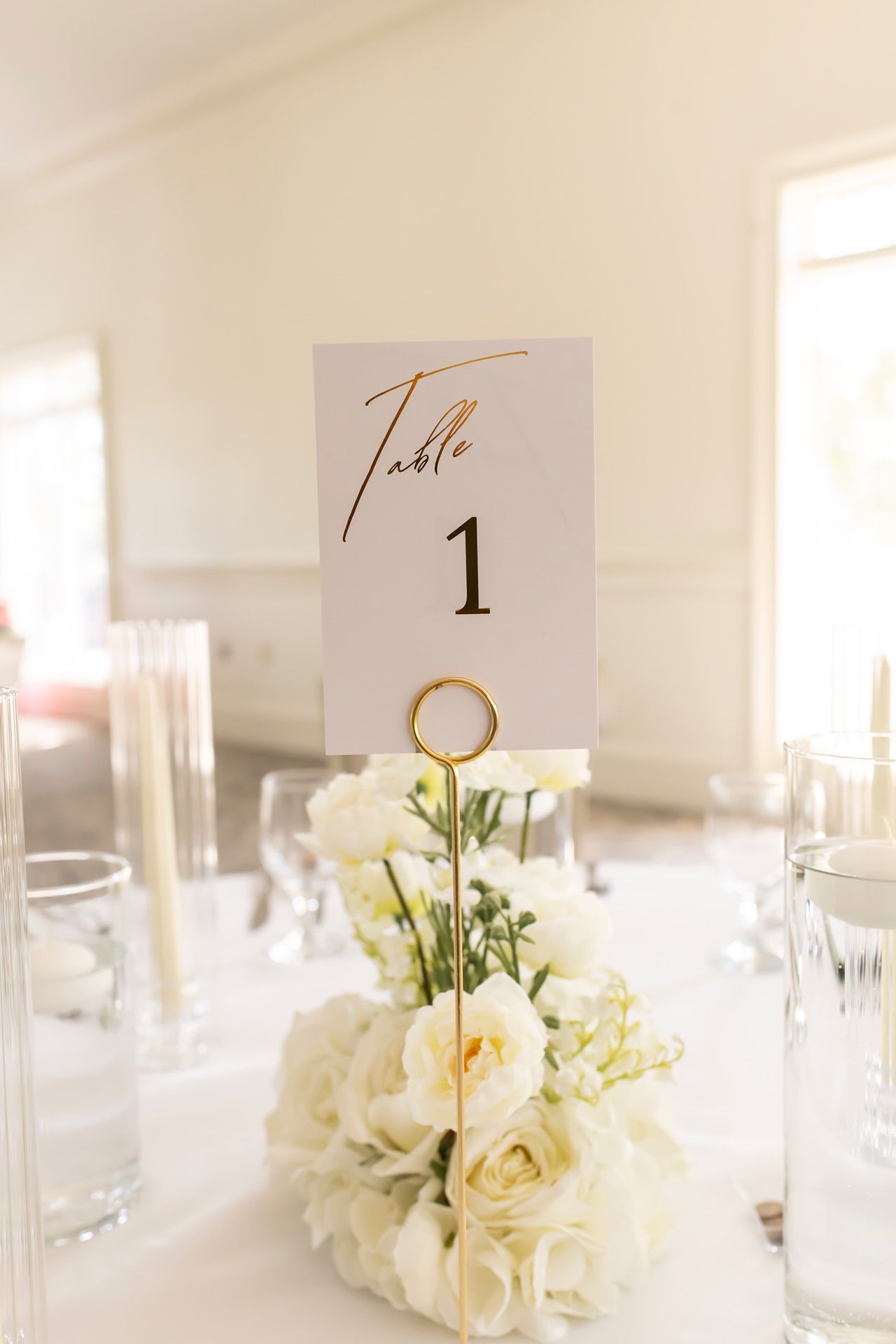 An elegant minimalist wedding table setting features white roses and clear vases with gold accented table number cards.