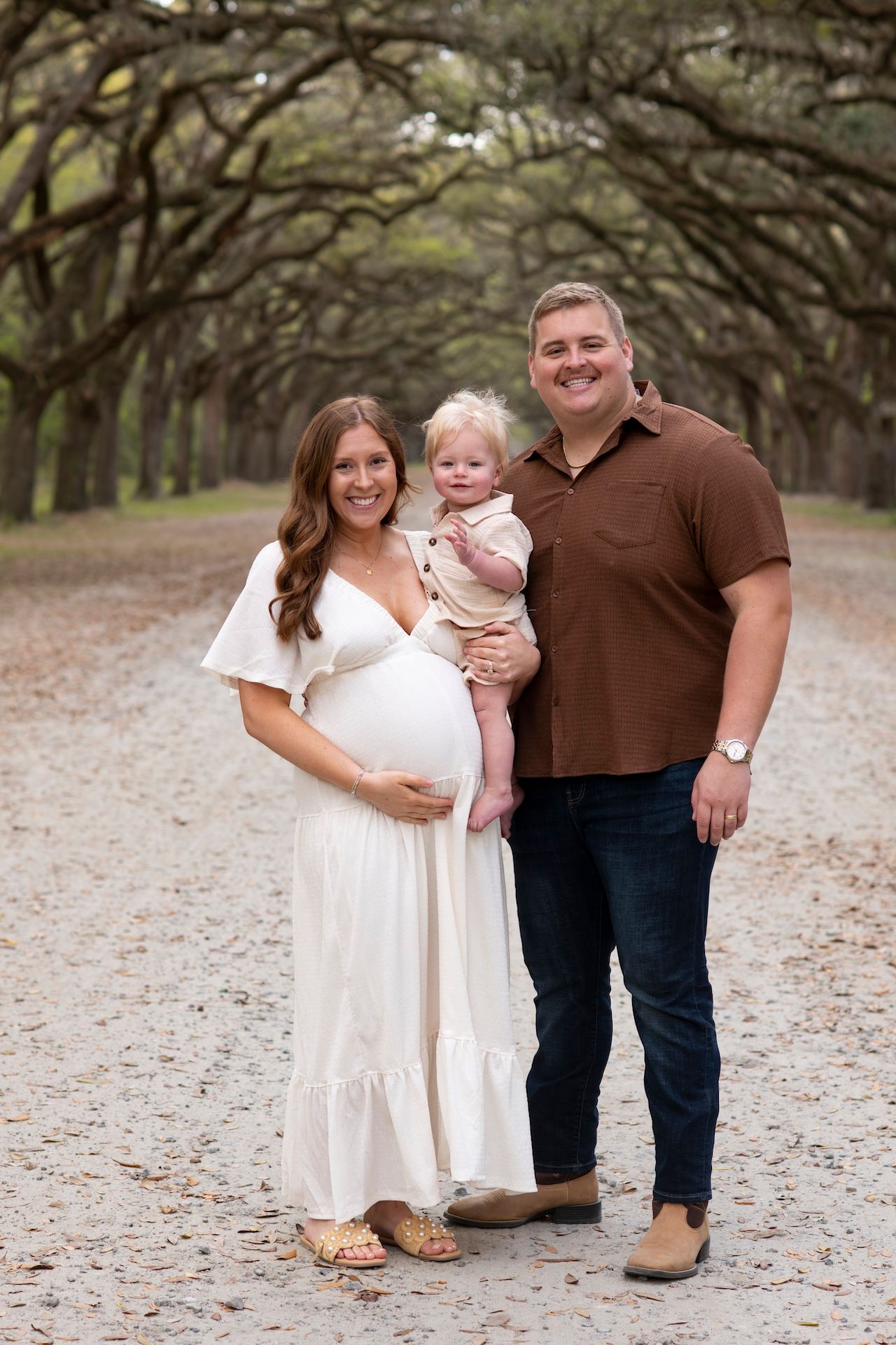 Pregnant woman in white dress poses with husband and toddler on a scenic tree-lined path for maternity photos.