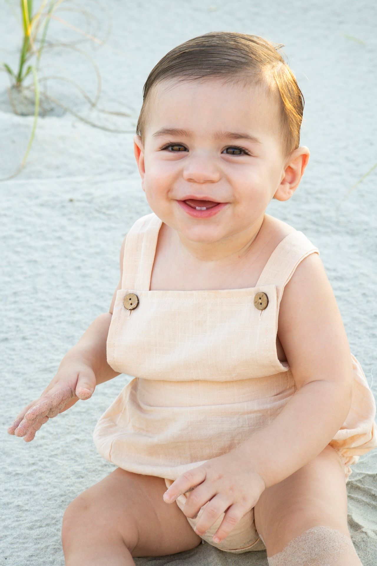 Smiling baby in a beige linen romper sitting on white sand at the beach, looking happy and cheerful.