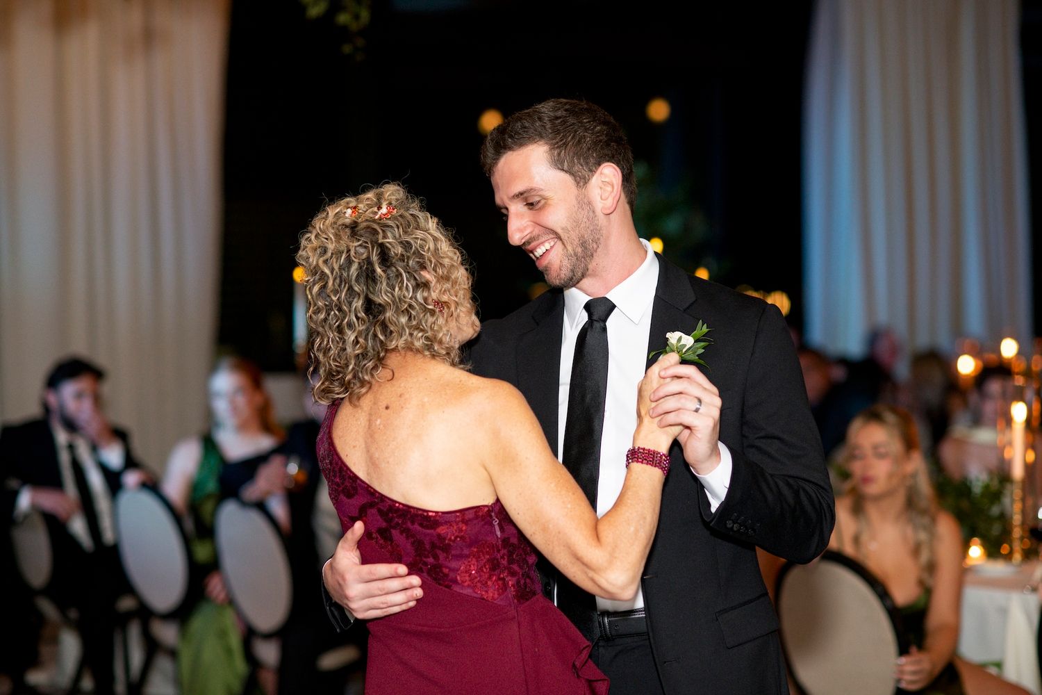 A couple dances together at an elegant evening wedding reception with soft lighting and other guests in the background.