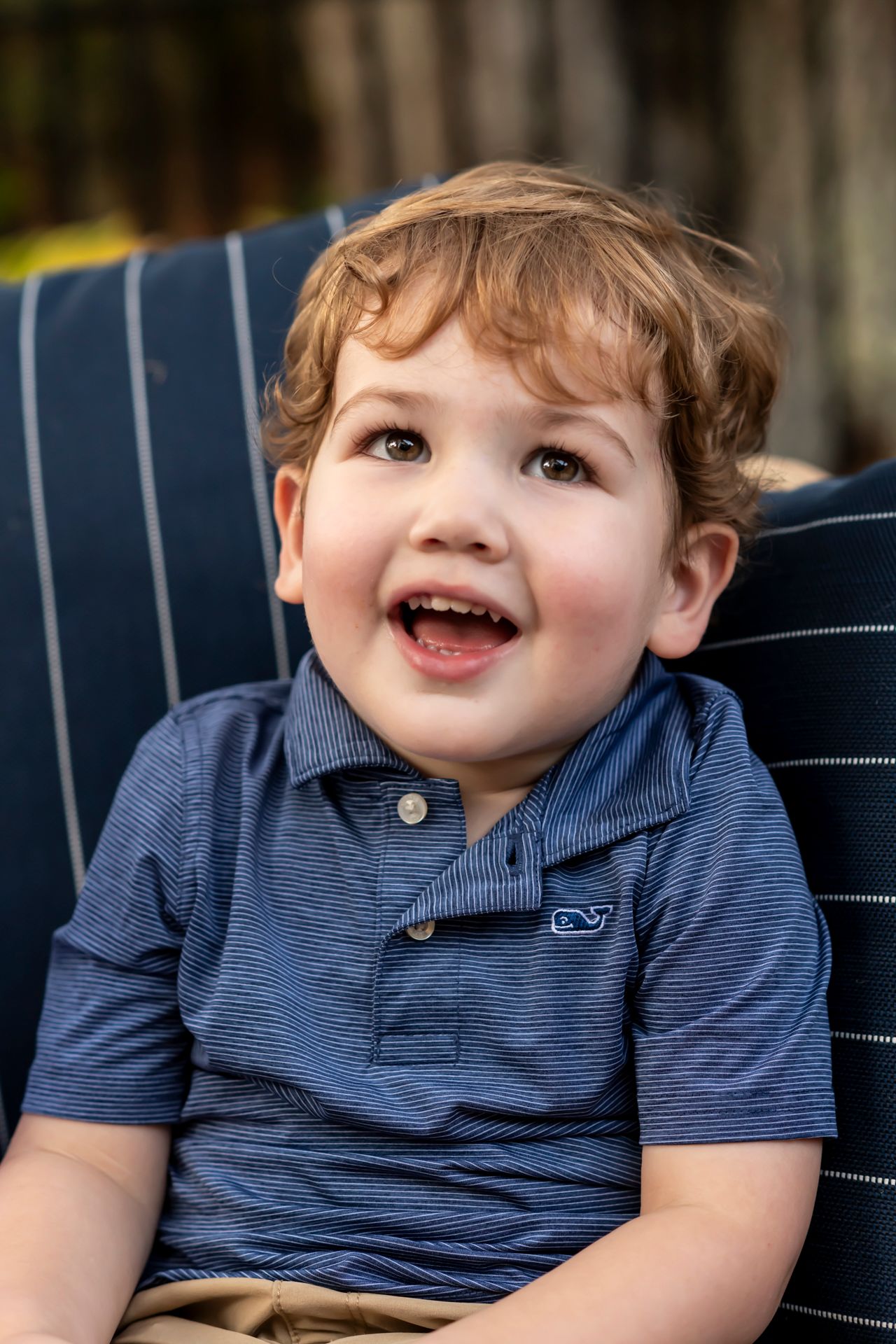 Young child in navy blue polo shirt sits on striped cushion while smiling and looking up with joy.