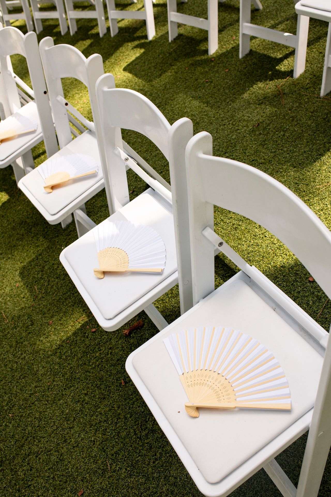 White folding chairs with decorative hand fans on seats arranged in rows on green grass for an outdoor wedding ceremony.