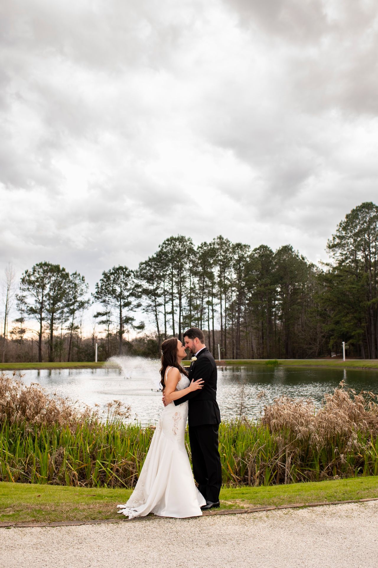 Bride and groom share a romantic kiss by a serene pond with tall pine trees and dramatic cloudy skies.