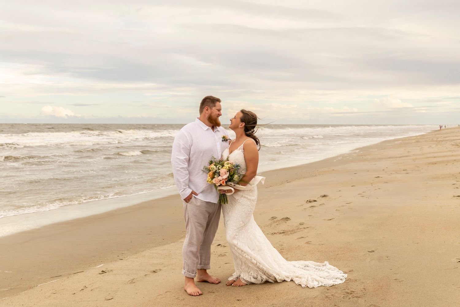 A couple in wedding attire embraces on a serene beach at sunset as waves roll onto the sandy shore.