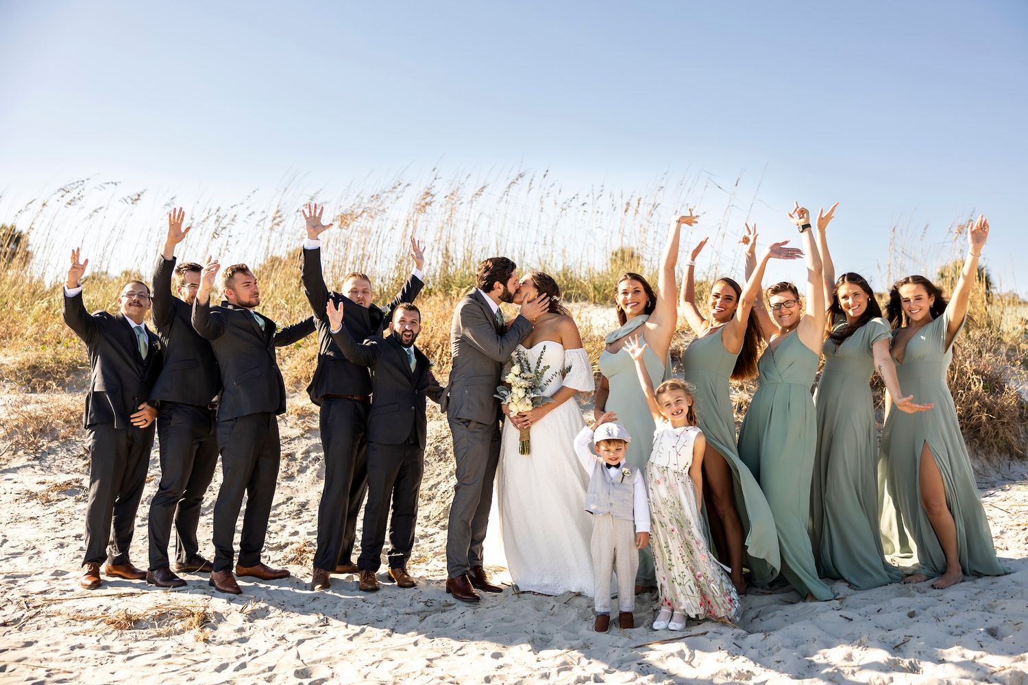 Wedding party celebrates joyfully with raised arms on snowy beach with tall grass in background.