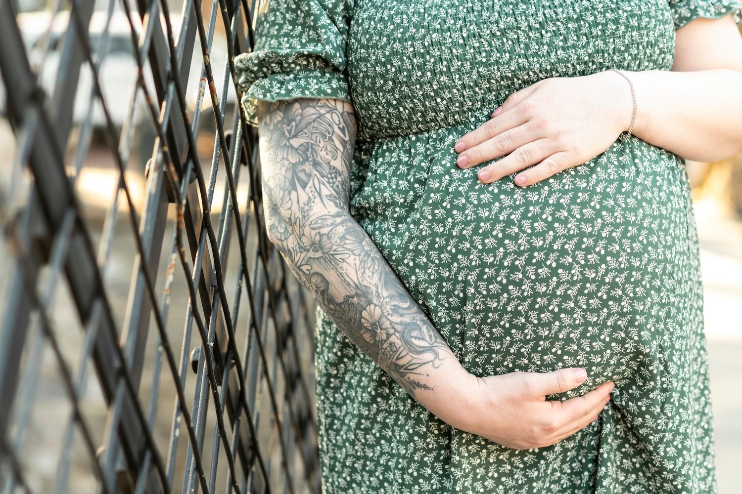 Close up of a pregnant belly in a green floral dress with tattooed arms cradling the bump against a lattice backdrop.