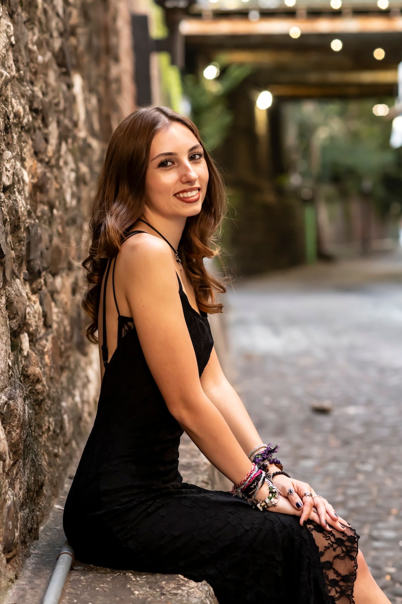 A woman in a black dress sits on a stone ledge along a cobblestone path with bokeh lights in the background.
