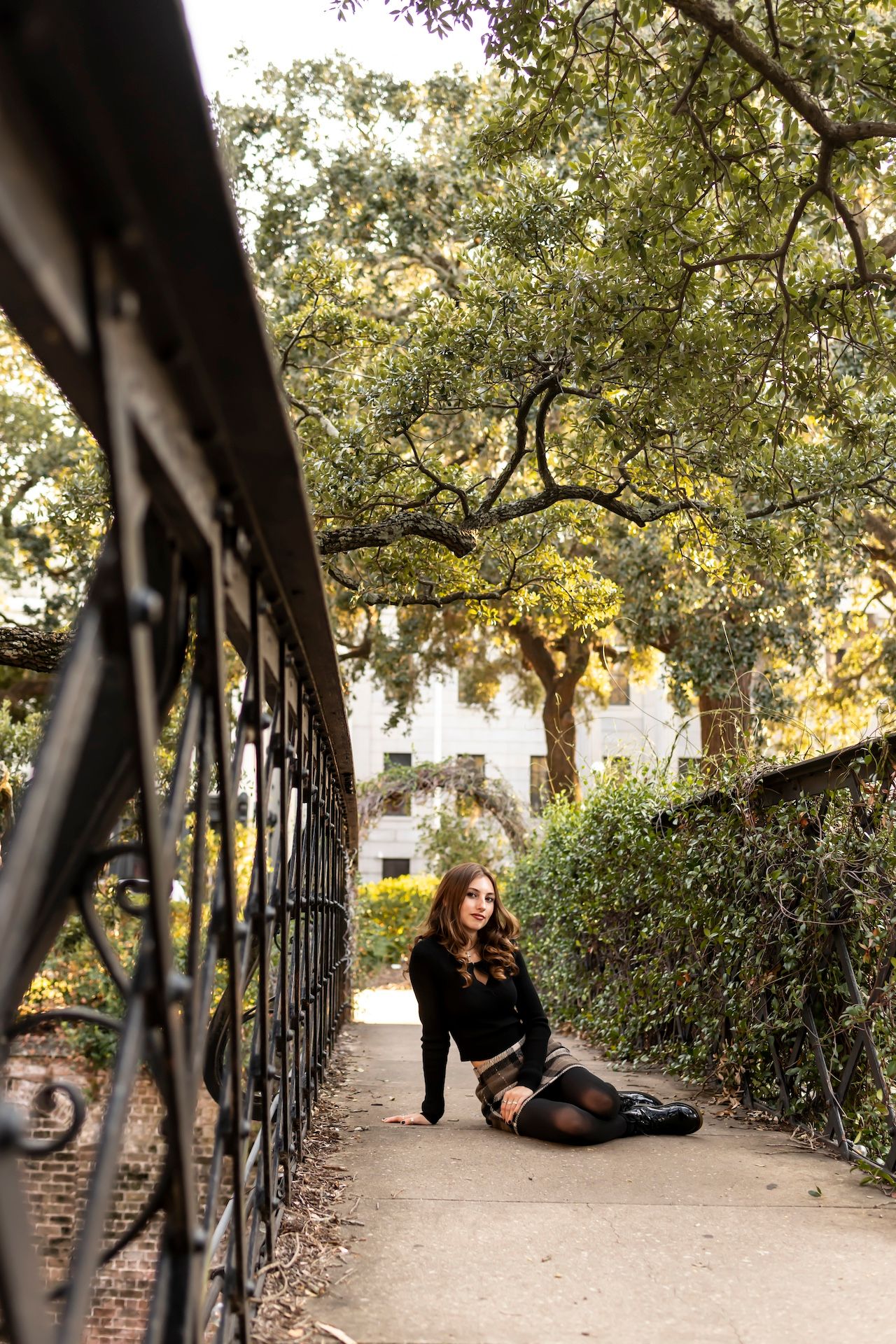 Someone in a black sweater poses on a brick pathway next to an ornate iron railing under oak trees in a historic setting.