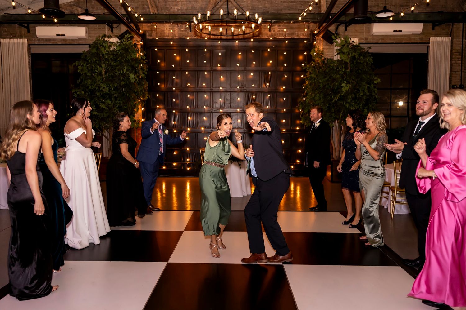 Wedding guests dance and celebrate on a black and white checkered dance floor under string lights.