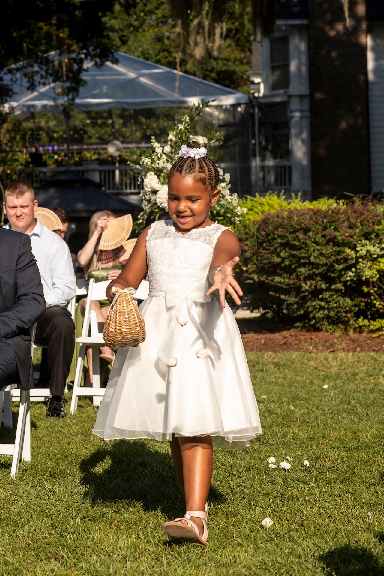 Small flower girls in white dresses playing and walking on grass during an outdoor wedding ceremony.