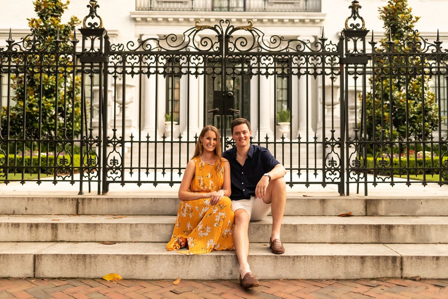 A couple shares a romantic moment together while sitting on stone steps in front of an ornate iron gate during golden hour.