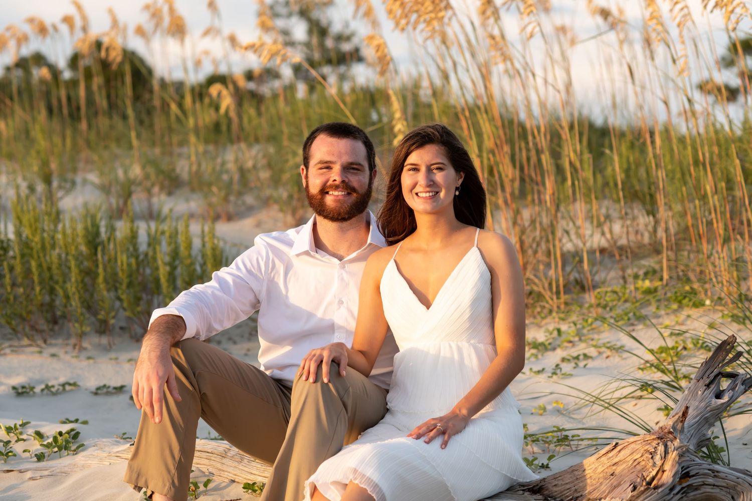 A couple in white attire sits together on a beach at sunset with sea oats in the background.