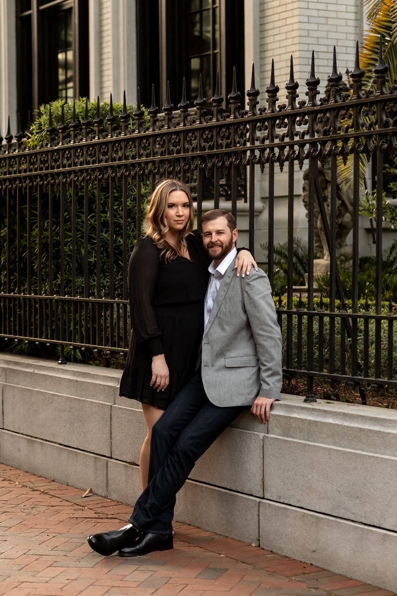 Couple poses together on brick sidewalk against wrought iron fence and stone wall in series of romantic engagement photos.